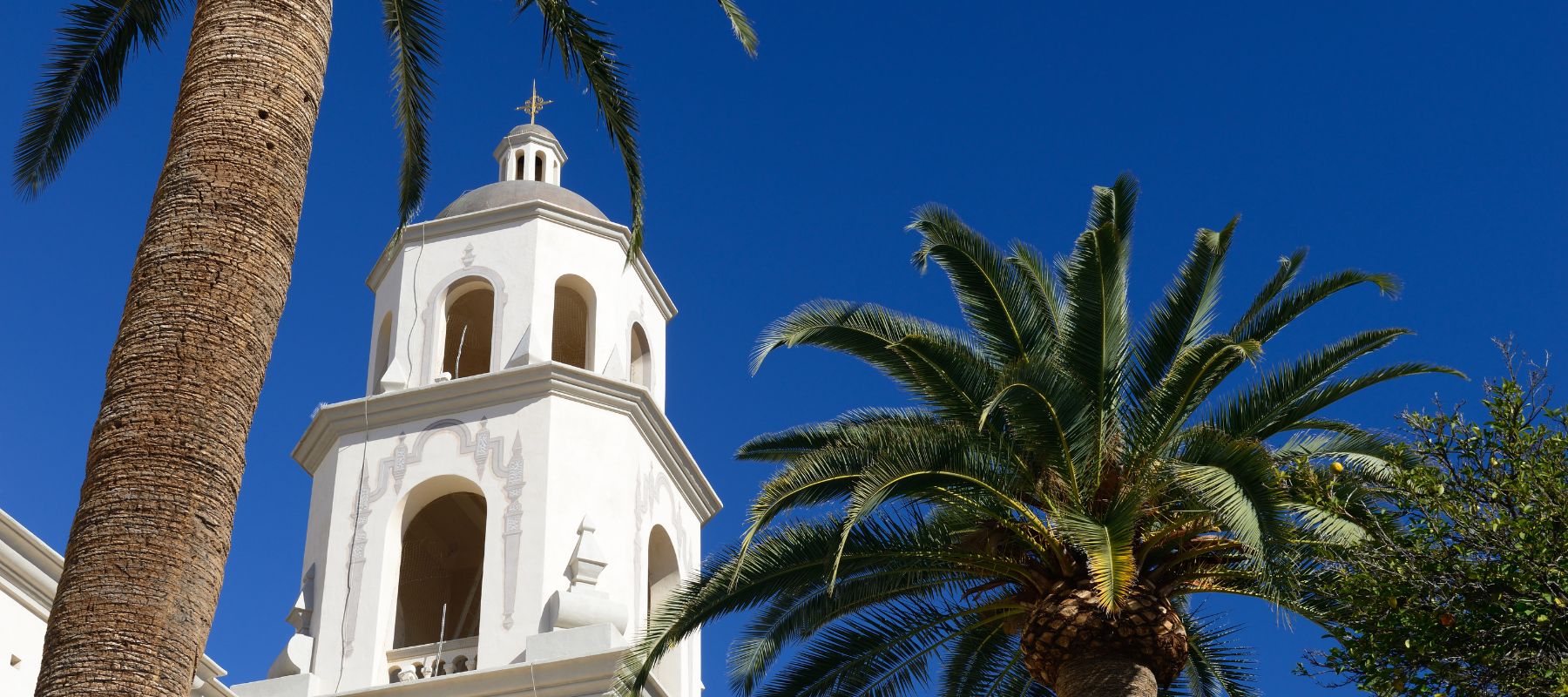 A picturesque white church tower with a red tiled roof stands under a clear blue sky, flanked by tall palm trees. The church's architecture features a traditional bell tower with visible arches and a cross at the top, typical of Spanish-style buildings found in warm, sunny climates.