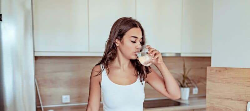woman in white tank top standing in the kitchen drinking a glass of water