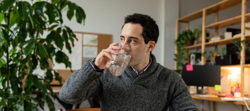 A man in a cozy office setting drinks a glass of water while sitting at his desk surrounded by plants and shelving