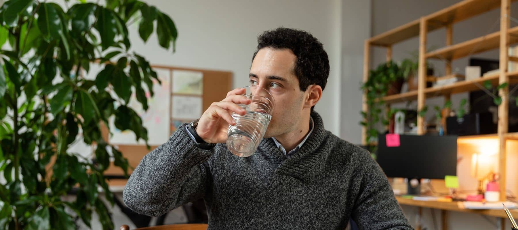 A man in a cozy office setting drinks a glass of water while sitting at his desk surrounded by plants and shelving